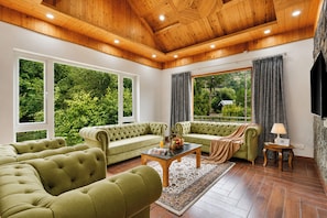 Un salón espacioso con sofá verde, una mesita de centro y un televisor. Amplias ventanas ofrecen vistas al exterior con vegetación.