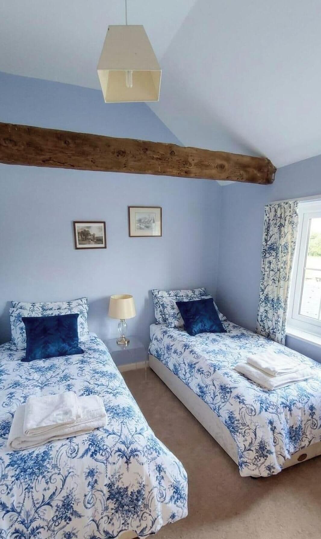 A bedroom with two beds, a wooden ceiling beam, a window with curtains, and two framed pictures on the wall.