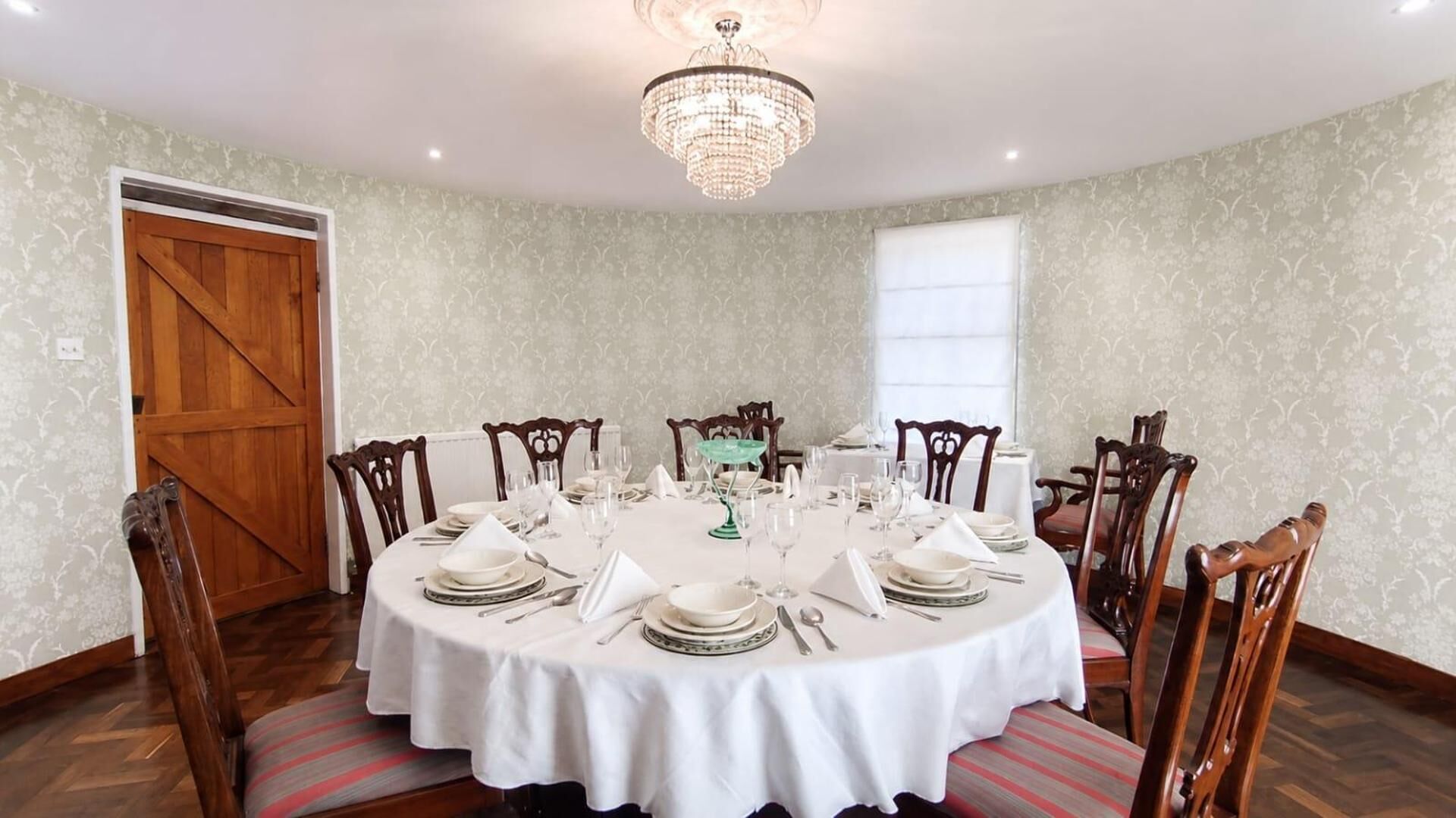 A dining room with a round table set for six, surrounded by wooden chairs, a chandelier, and patterned wallpaper.