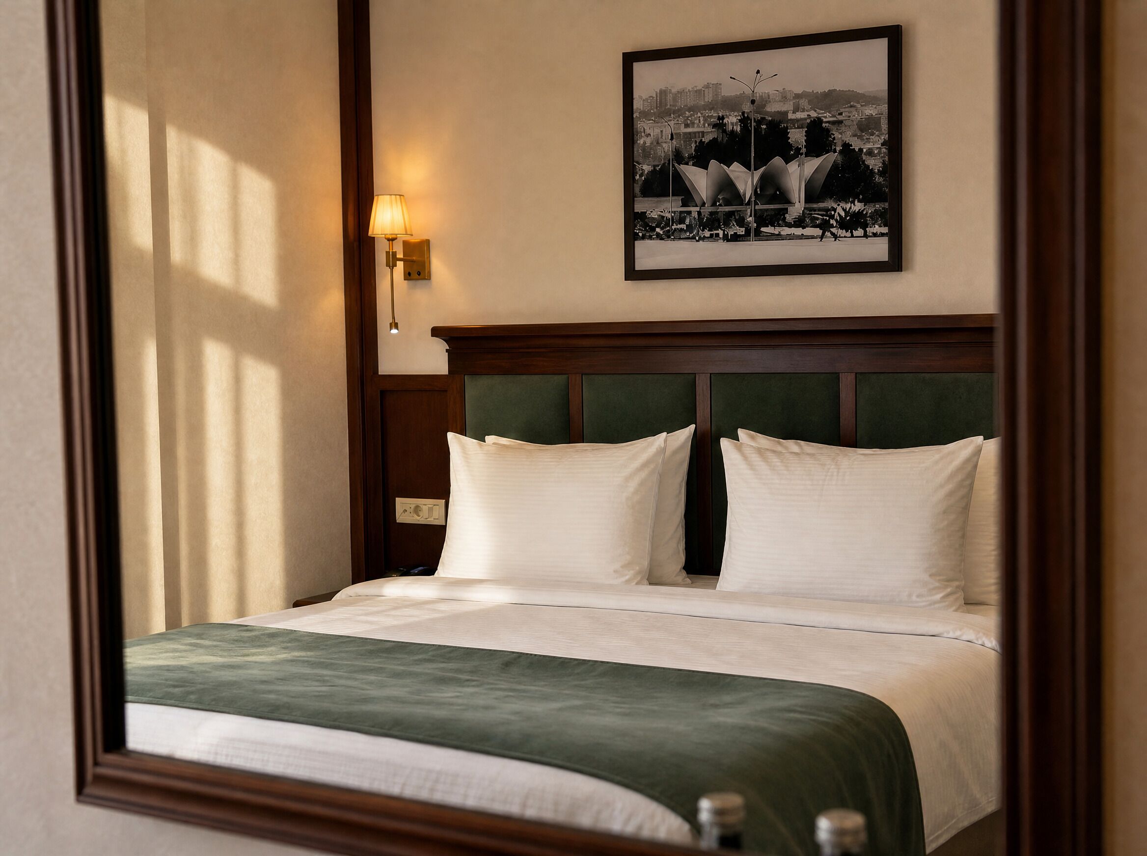 A neatly made bed with a green throw, a wall-mounted lamp, and a framed cityscape picture above it.