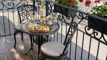 Una mesa redonda con una botella de vino, copas y una tabla de quesos en un balcón con barandas metálicas y macetas de flores rojas.