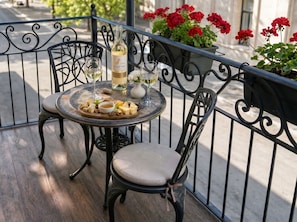 Een ronde tafel met een fles wijn, glazen en een kaasplank op een balkon met metalen hekwerk en rode bloempotten.
