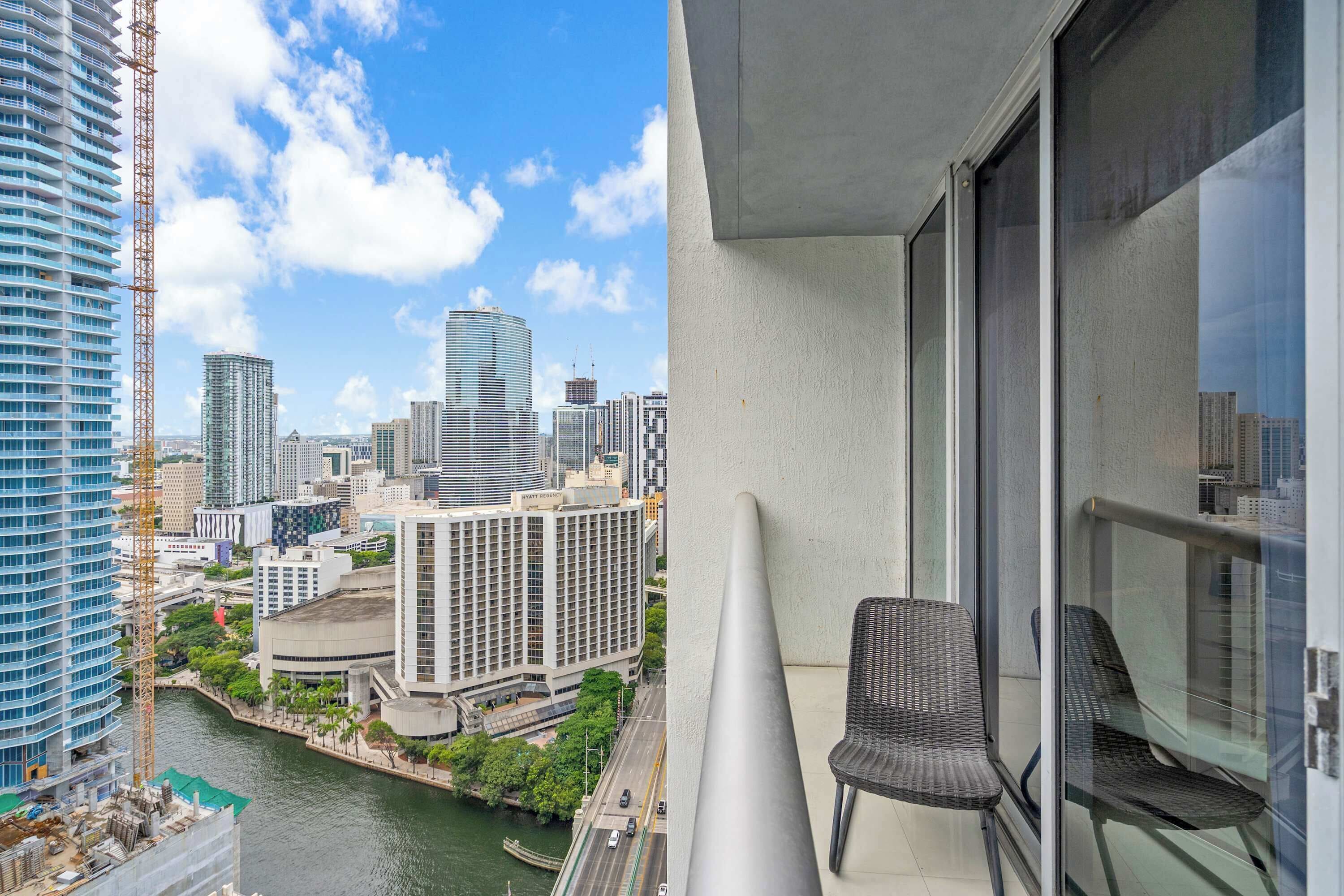 A high-rise building with a view of a city skyline, a balcony with a chair, and a river.
