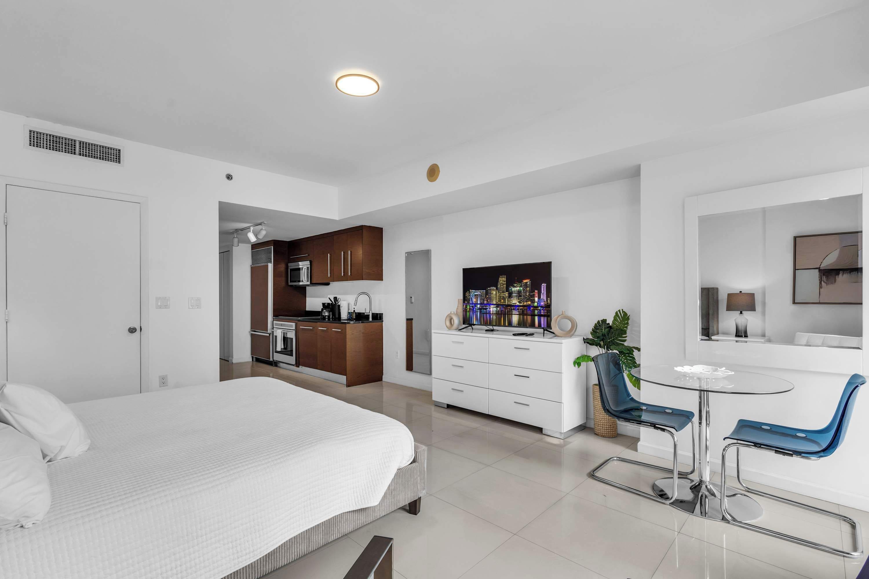 A modern hotel room with a bed, a kitchenette, a dining area with a table and chairs, and a TV.