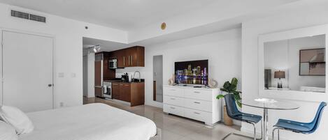 A modern hotel room with a bed, a kitchenette, a dining area with a table and chairs, and a TV.