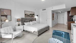 A modern bedroom with a bed, bedside tables, a chair, a painting, and a view into the kitchen. - Luxe Condo on Miami River (Miami)