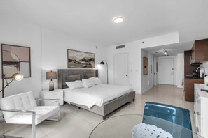 A modern bedroom with a bed, bedside tables, a chair, a painting, and a view into the kitchen. - Luxe Condo on Miami River (Miami)