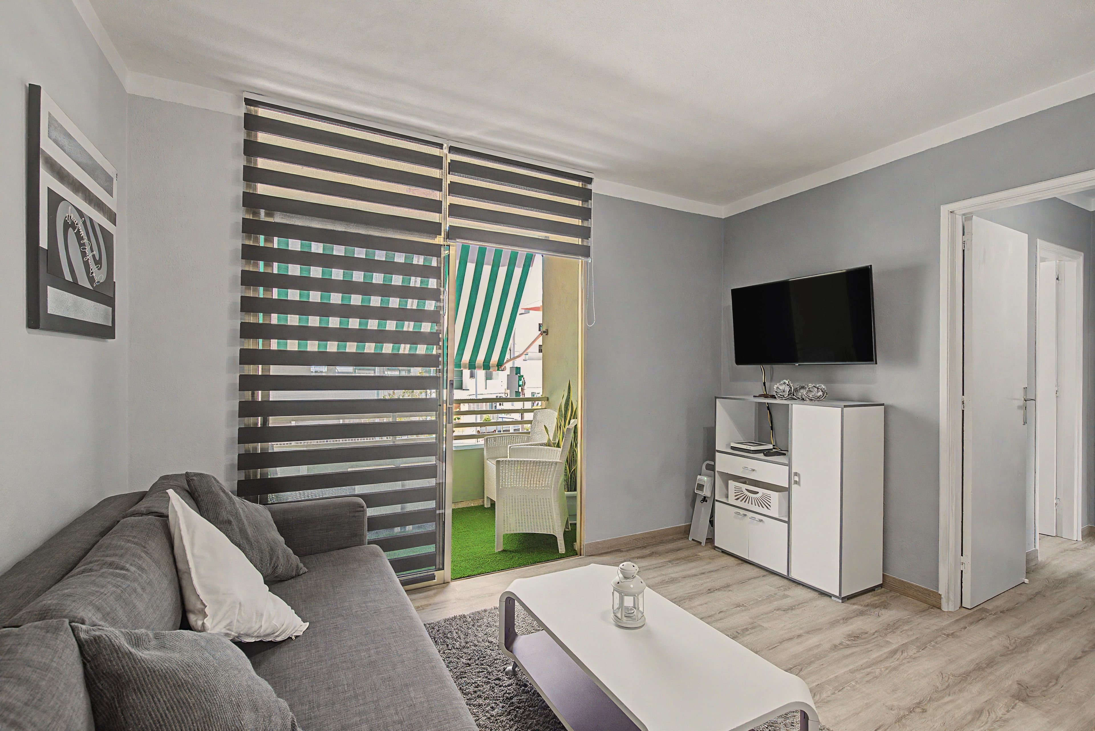Una sala de estar moderna con un sofá gris, una mesa de centro blanca, un televisor de pantalla plana y un puertas correderas que dan a un balcón.