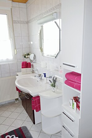 A modern bathroom with a white sink, mirror, and white tiles. There is a window with a view of the outside, a radiator, and a towel rack with pink towels. - SeeZeit Dingelsdorf - Modern Vacation Rental with Lake Views and Access to Bicycle Trails (Konstanz)