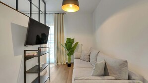 A modern living room with a flat-screen TV, a black shelving unit, a grey sofa, and a large potted plant. - Dwie Motławy 59 | Riverside | Perfect Stay (Gdańsk)