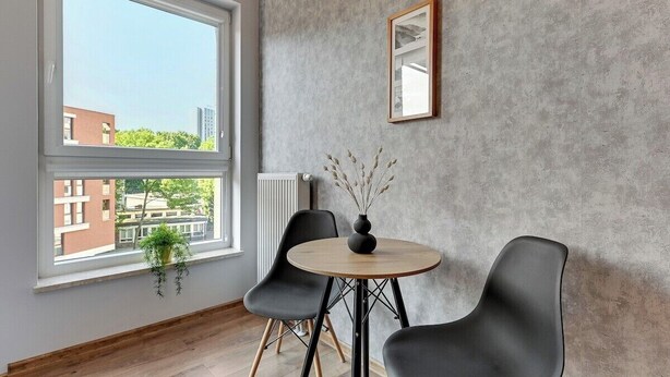 Una habitación con un ventanal grande, una mesa redonda con sillas, una planta en maceta y un cuadro colgado en la pared.