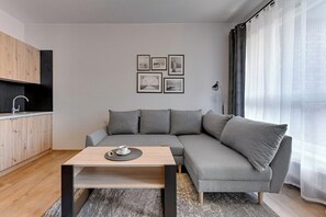 A modern living room with a grey sectional sofa, a wooden coffee table, and a kitchen area with a black cabinet and a sink. - City Center | Bastion Wałowa 59 with Balcony (Gdańsk)