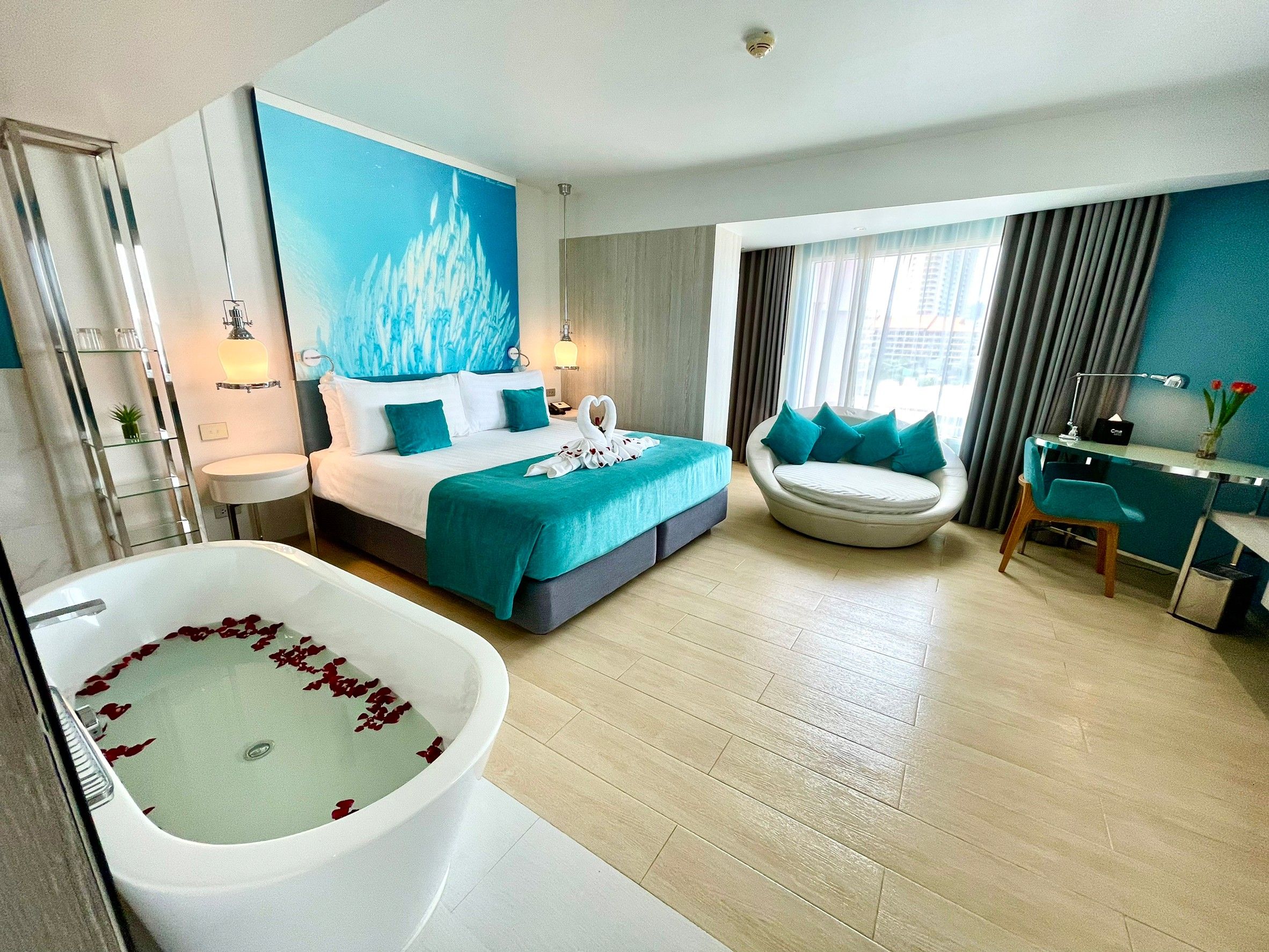 A modern hotel room with a large bed, a separate seating area, a bathtub with rose petals, and a view of the city.