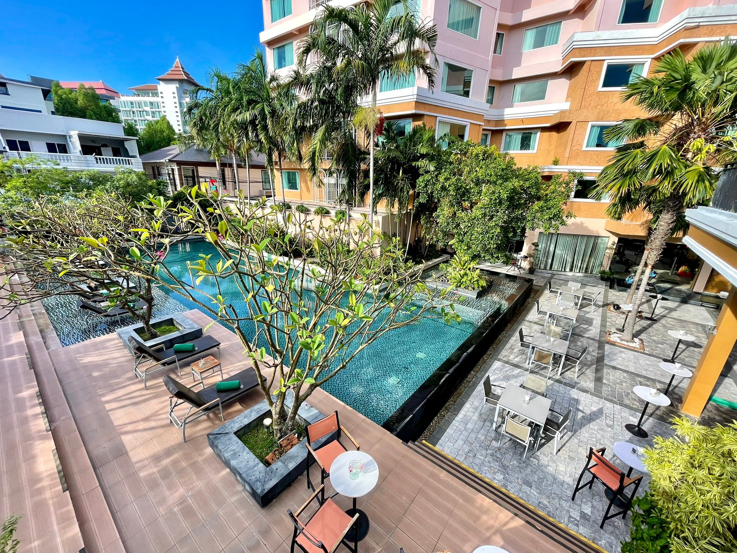 A rooftop pool area with lounge chairs and tables, surrounded by greenery and buildings.