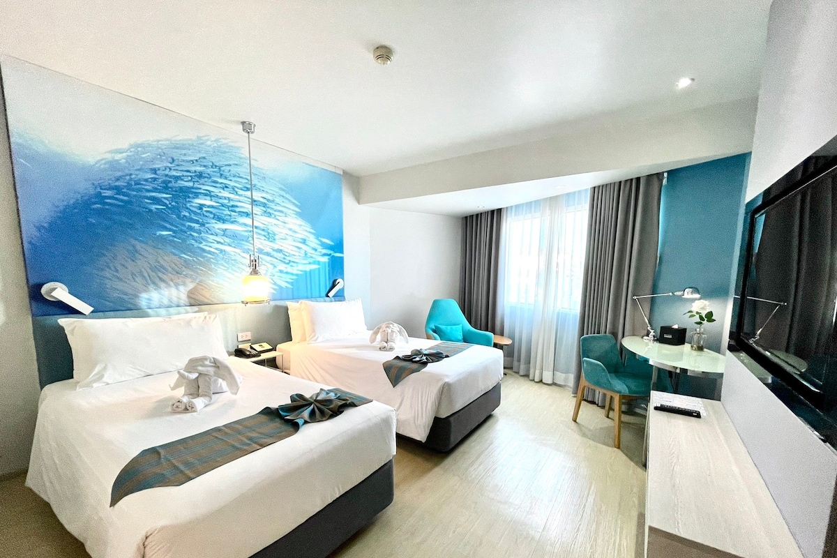 A hotel room with two beds, a desk, a chair, a TV, and a painting of a fish school on the wall.