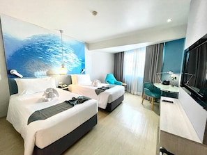 A hotel room with two beds, a desk, a chair, a TV, and a painting of a fish school on the wall. - J Tower Pattaya (Pattaya)