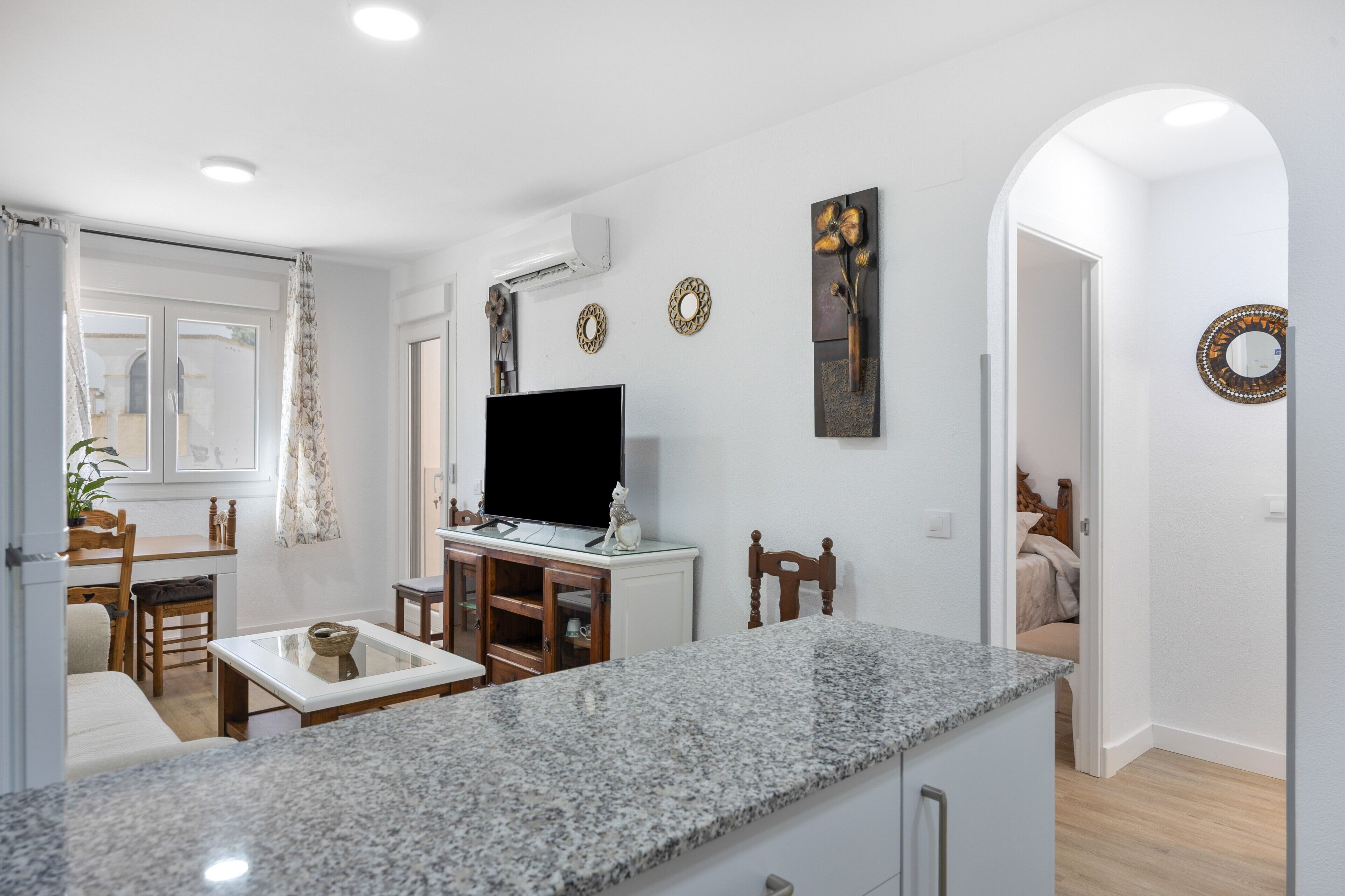 A modern kitchen with a granite countertop, a flat-screen TV, and a dining area with wooden chairs and a table.