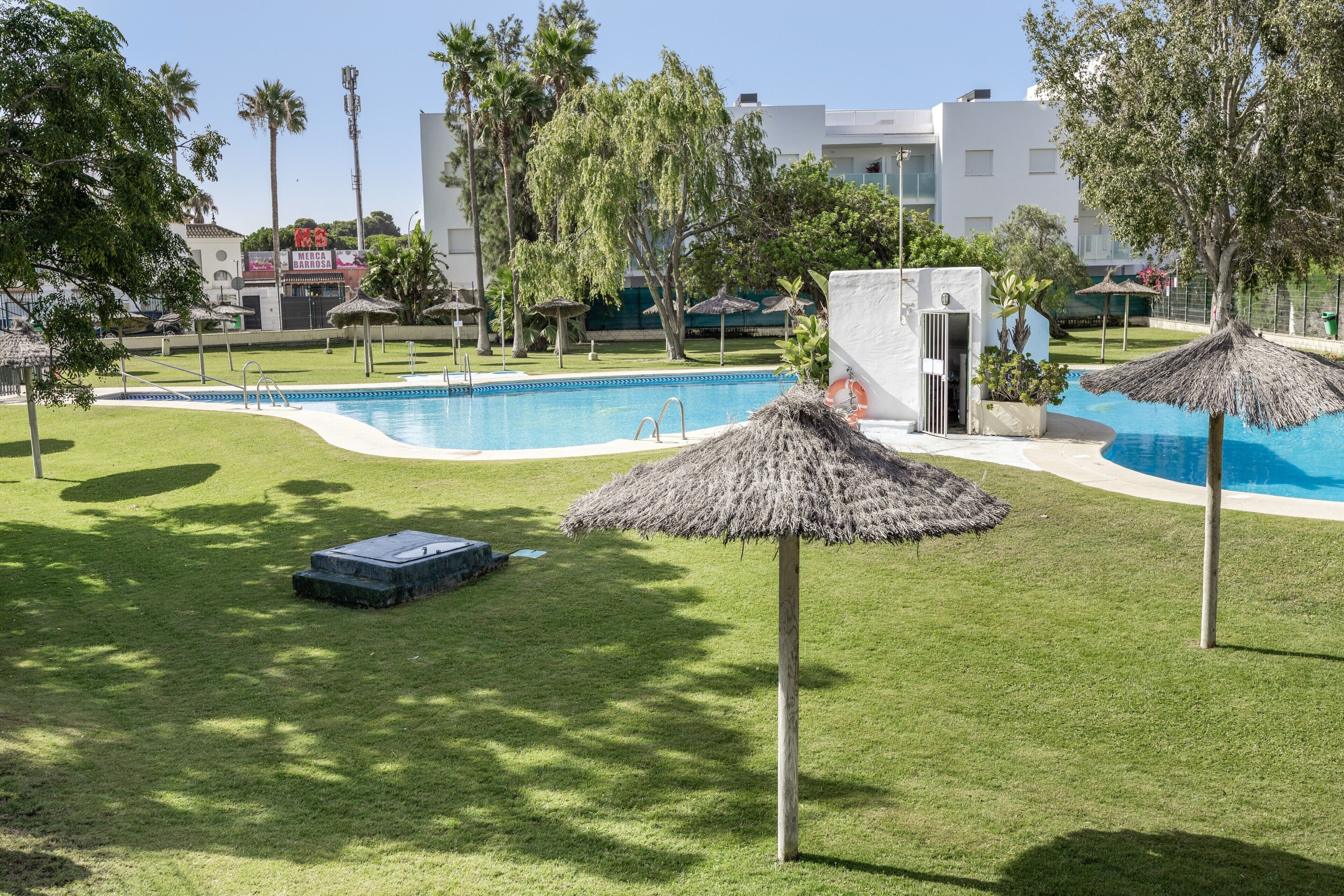 Una piscina con sombrilla de paja, rodeada de césped cuidado y árboles.