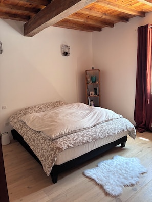 A bedroom with a bed, a wooden floor, a white rug, a shelf with books and decorative items, and a window with curtains. - Country house 45 minutes from Toulouse. Pets Allowed (Saint-Urcisse)