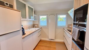 Una cuina compacta amb armaris blancs, un forn de mur i dues finestres que deixen entrar llum natural.