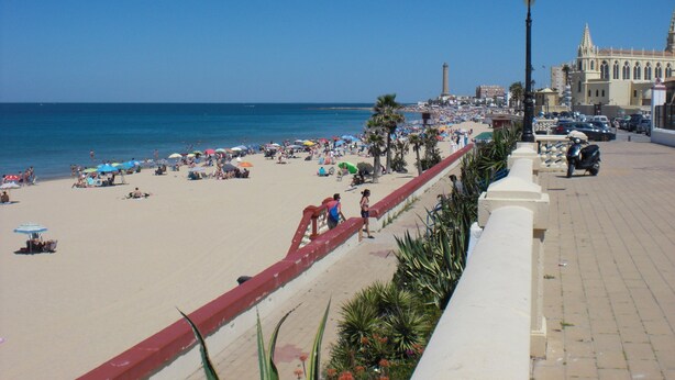 A beach with a paved walkway, palm trees, and a historic building in the background. - Apartamento Caracola (Chipiona)
