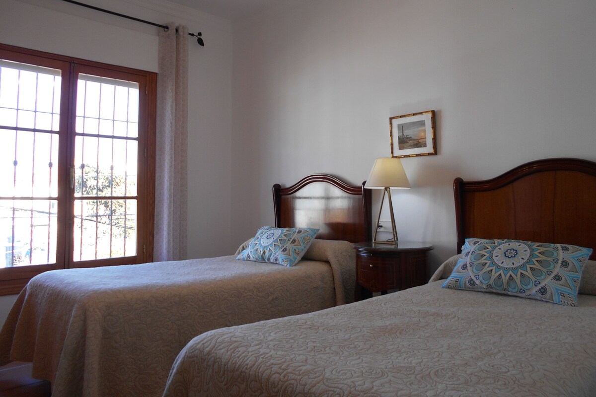 A room with two beds, a nightstand, a lamp, a picture on the wall, and a window with curtains.