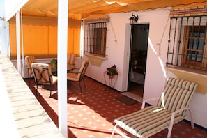 A covered outdoor seating area with chairs and a table, a door leading to an interior space, and a window with bars. - Apartamento Caracola (Chipiona)