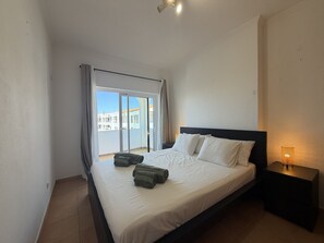 A hotel room with a bed, a nightstand, a lamp, and a view of buildings through a window. - Gale Beach Walk T3 Apartment (Albufeira)
