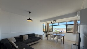 Una sala de estar moderna con un sofá modular, una mesa de comedor y sillas. Hay un televisor sobre un mueble bajo y una ventana con vistas a edificios.