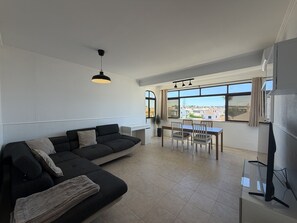 A modern living room with a sectional sofa, dining table, and chairs. There is a TV on a stand and a window with a view of buildings. - Gale Beach Walk T3 Apartment (Albufeira)