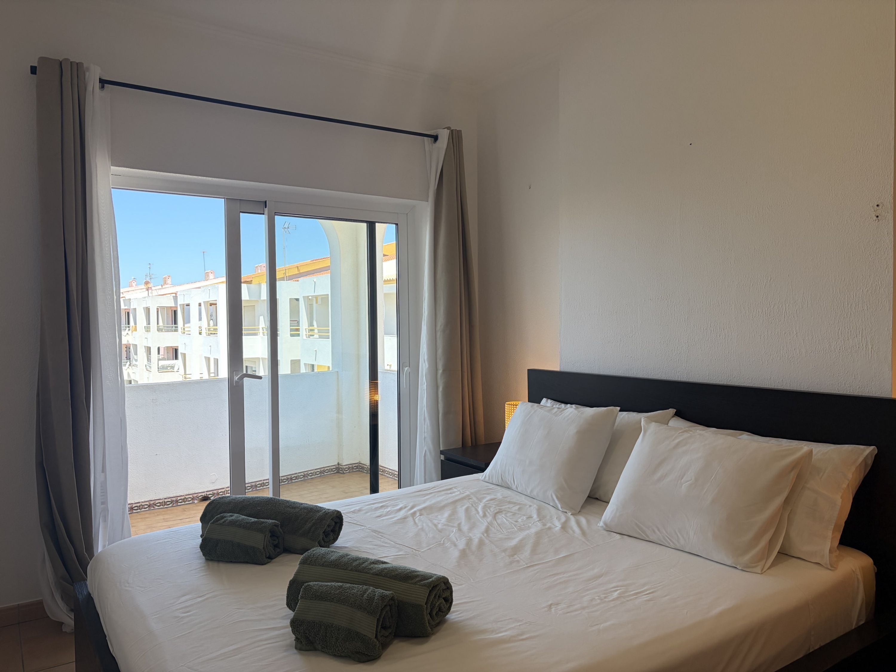 A hotel room with a bed, two pillows, a nightstand, and a view of buildings through a glass door.