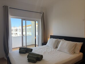 A hotel room with a bed, two pillows, a nightstand, and a view of buildings through a glass door. - Gale Beach Walk T3 Apartment (Albufeira)