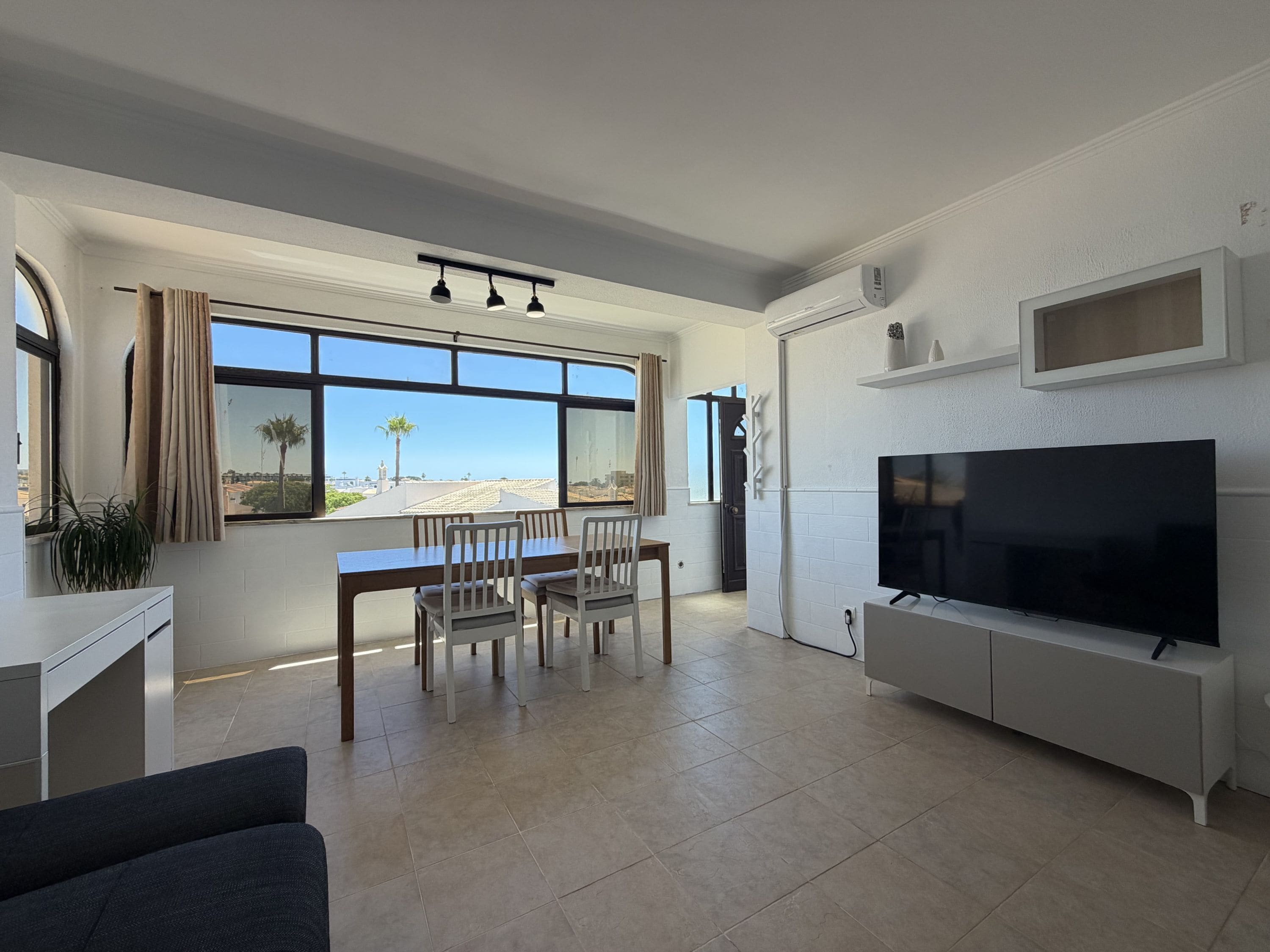 A modern living room with a dining area, a flat-screen TV, and a large window offering a view of the beach.