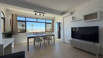 A modern living room with a dining area, a flat-screen TV, and a large window offering a view of the beach.