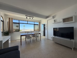 A modern living room with a dining area, a flat-screen TV, and a large window offering a view of the beach. - Gale Beach Walk T3 Apartment (Albufeira)
