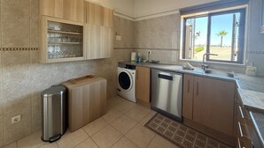 Una cocina moderna con lavadora integrada, un cubo de basura de acero inoxidable y una ventana con vista a palmeras.