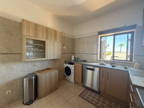 A modern kitchen with a built-in washing machine, a stainless steel trash bin, and a window with a view of palm trees. - Gale Beach Walk T3 Apartment (Albufeira)