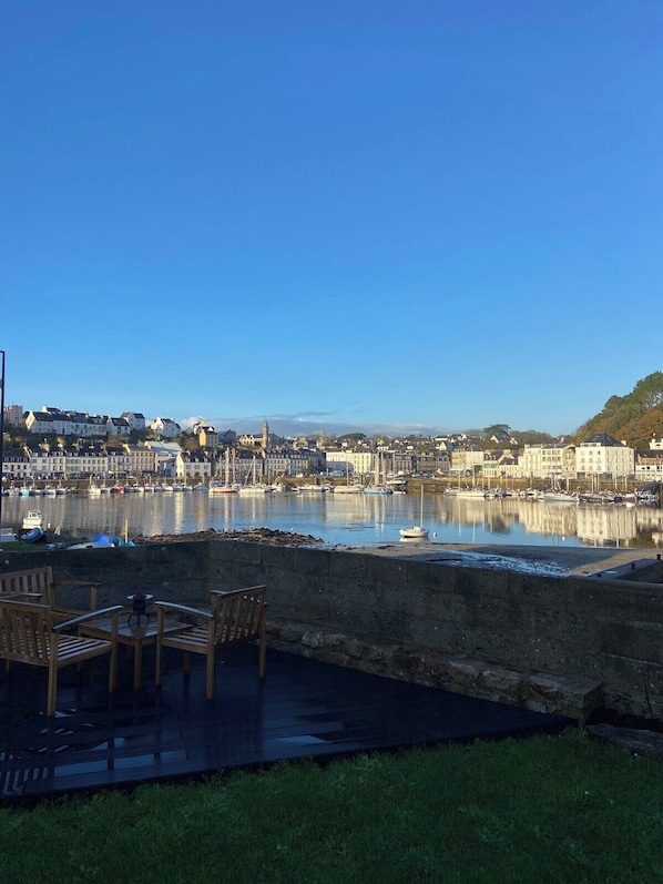 A waterfront view with a row of buildings, a calm river, and a grassy area with chairs. - A garden on the beautiful port of Audierne (Plouhinec)