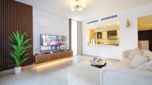 A modern living room with a flat-screen TV, a sofa, a potted plant, and a kitchen area visible through an open door. - Modern Home w/ Rooftop Terrace in Prime Location (Al Hamriyah)