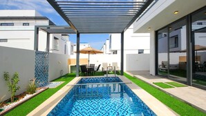 A modern outdoor swimming pool with a glass ceiling, surrounded by a patio area with seating and a view of the cityscape. - Modern Home w/ Rooftop Terrace in Prime Location (Al Hamriyah)