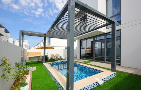 A modern outdoor pool area with a glass enclosure, artificial grass, and a pergola structure.