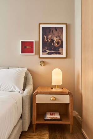 A neatly arranged hotel room with a bed, a bedside table with a lamp, a framed picture, and a small shelf with a book. - Close to American Airlines Center + Dining & Pool (Dallas)