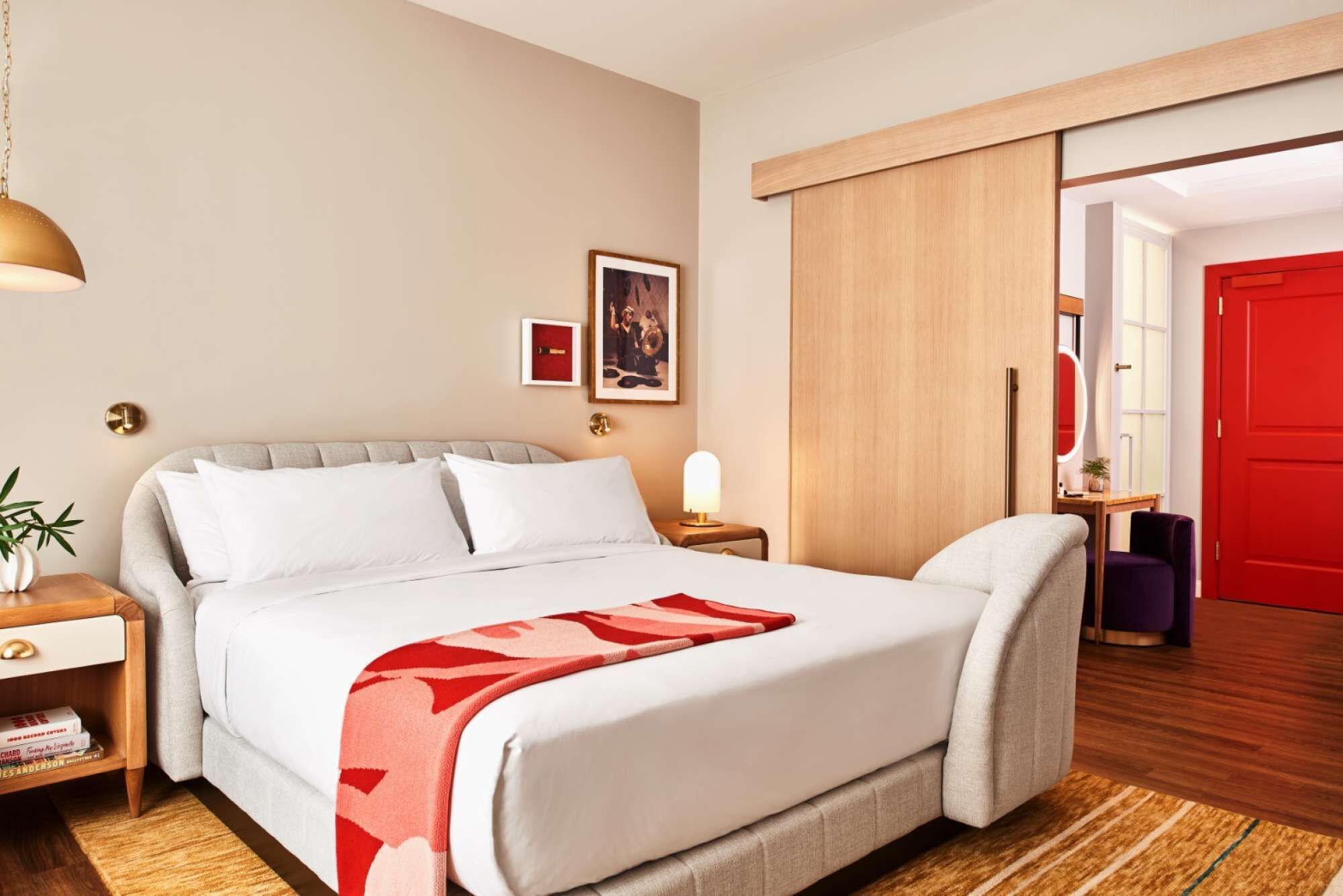 A hotel room with a bed, bedside tables, a lamp, a red door, and a view into another room.