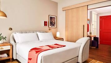 A hotel room with a bed, bedside tables, a lamp, a red door, and a view into another room.