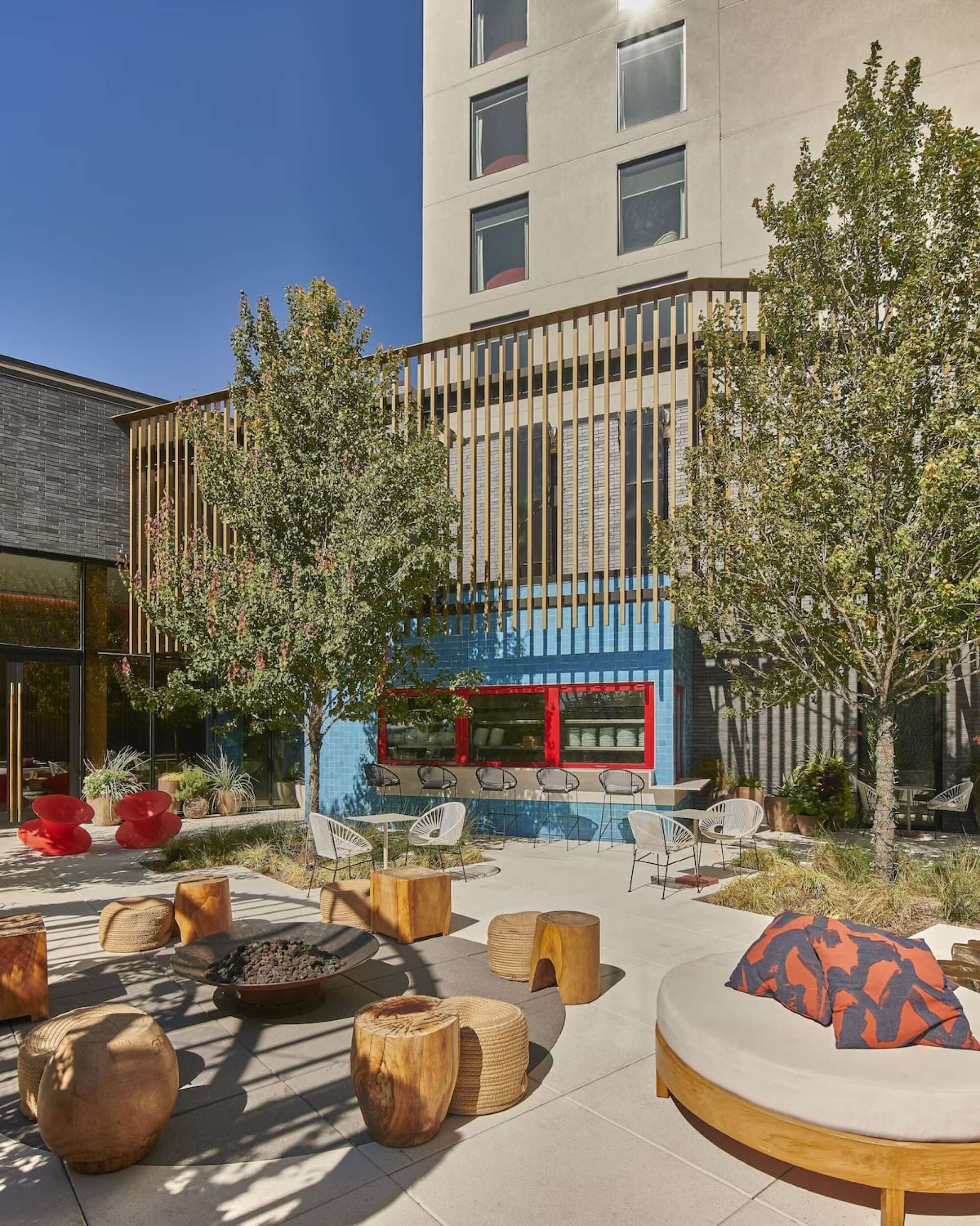 An outdoor seating area with round wooden tables and chairs, a fire pit, and a building with a blue facade in the background.