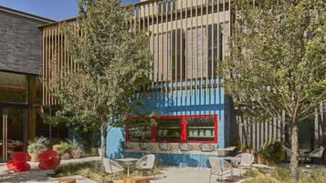 An outdoor seating area with round wooden tables and chairs, a fire pit, and a building with a blue facade in the background.