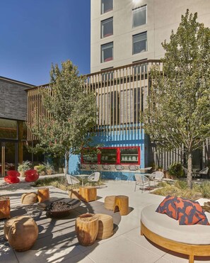 An outdoor seating area with round wooden tables and chairs, a fire pit, and a building with a blue facade in the background. - Dallas' Design District Views + Rooftop Pool & Bar (Dallas)