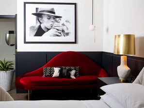 A red sofa with patterned pillows, a framed black and white photograph on the wall, and a gold lamp on a bedside table. - Dallas' Design District Views + Rooftop Pool & Bar (Dallas)