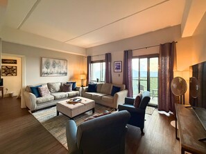 Ein modernes Wohnzimmer mit einer Sofagarnitur, einem Couchtisch und einem Fernseher. Zwei Fenster mit Vorhängen und ein durch die Fenster sichtbarer Balkon.