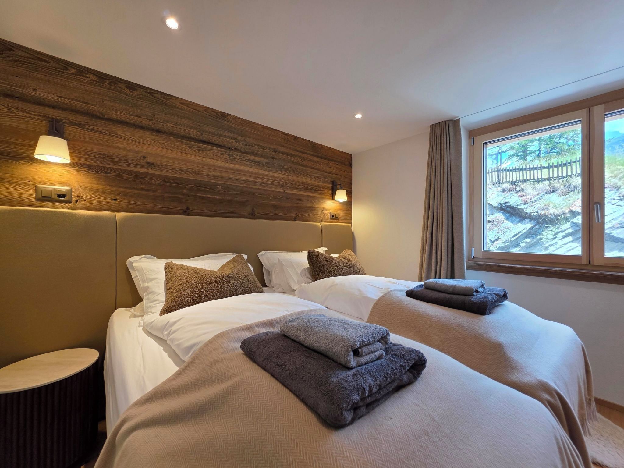 A hotel room with a wooden headboard, two beds with neatly made beds and towels, a round side table, and a window with a view of a fence and trees.